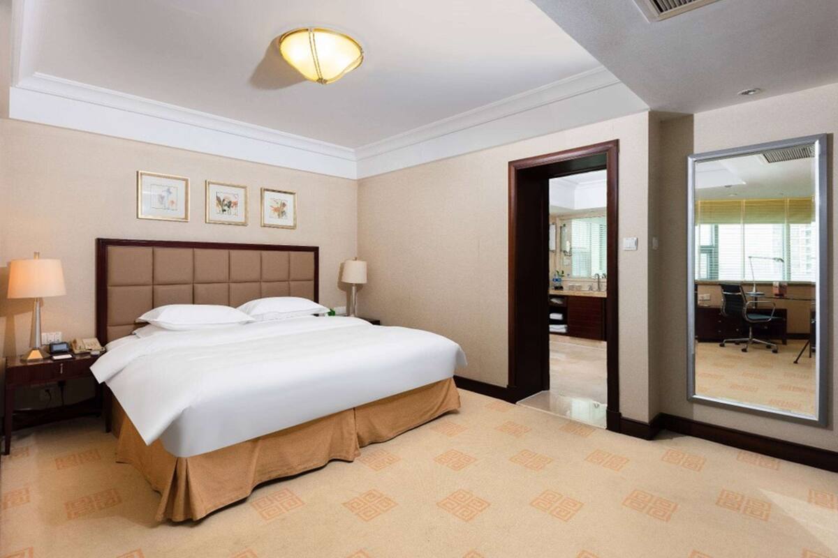 A hotel room with a large bed, a bedside table with a lamp, a mirror, and framed pictures on the wall.