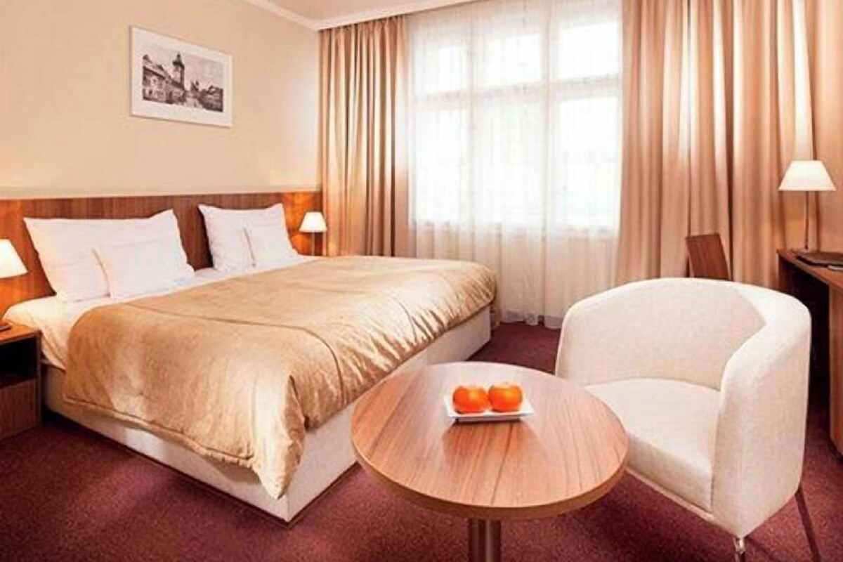A hotel room with a bed, a round table with oranges, a white armchair, and a picture on the wall.