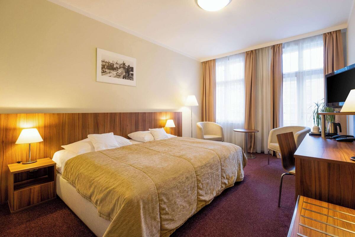 Een hotelkamer met een groot bed, twee nachtkastjes met lampen, een bureau met stoel, een televisie en een raam met gordijnen.