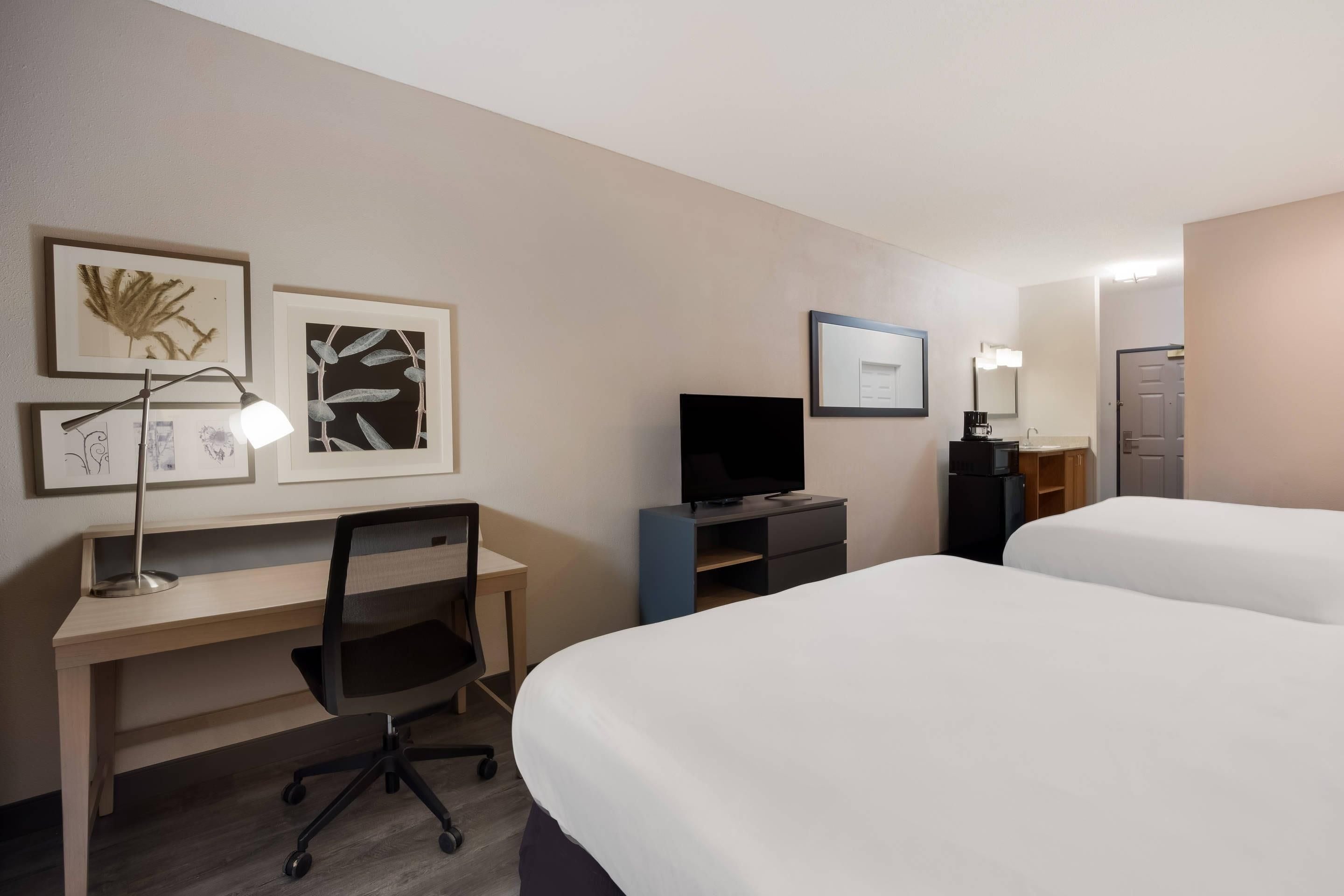 Una habitación de hotel con escritorio, silla, televisor, dos camas y obras de arte en las paredes.