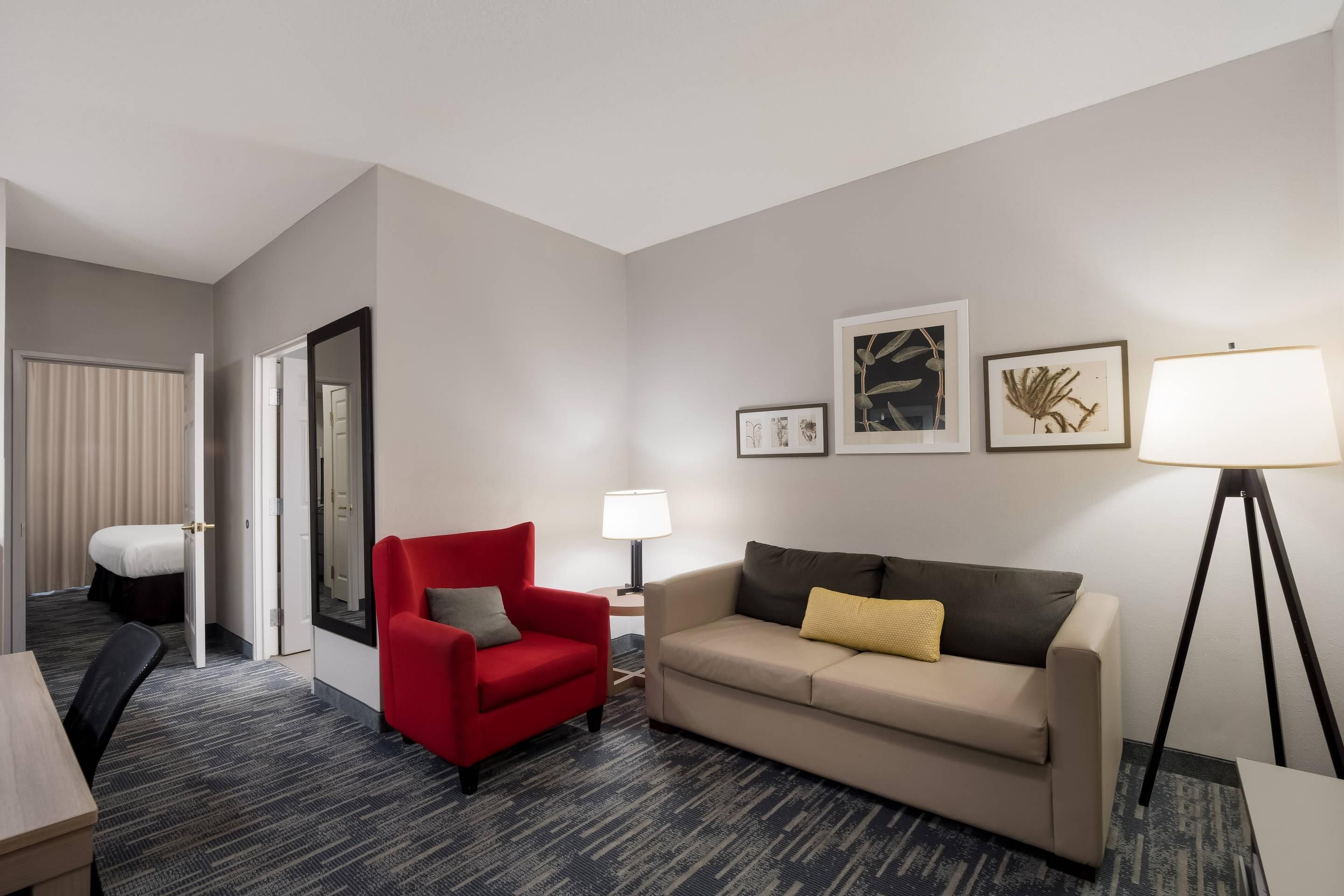 A hotel room with a red armchair, a beige sofa, a floor lamp, and framed botanical prints on the wall.