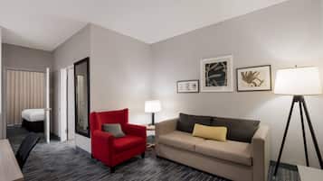 A hotel room with a red armchair, a beige sofa, a floor lamp, and framed botanical prints on the wall.