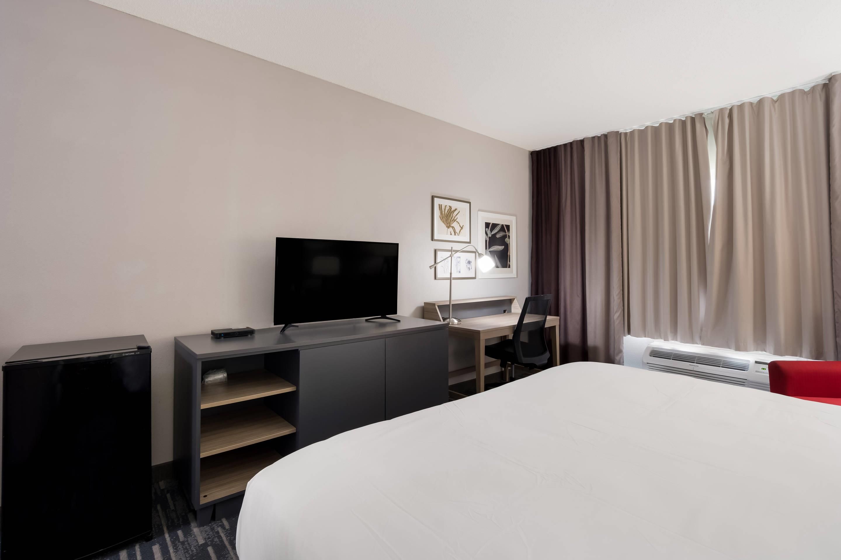 Habitación de hotel con cama, escritorio, televisor, silla y ventana con cortinas.