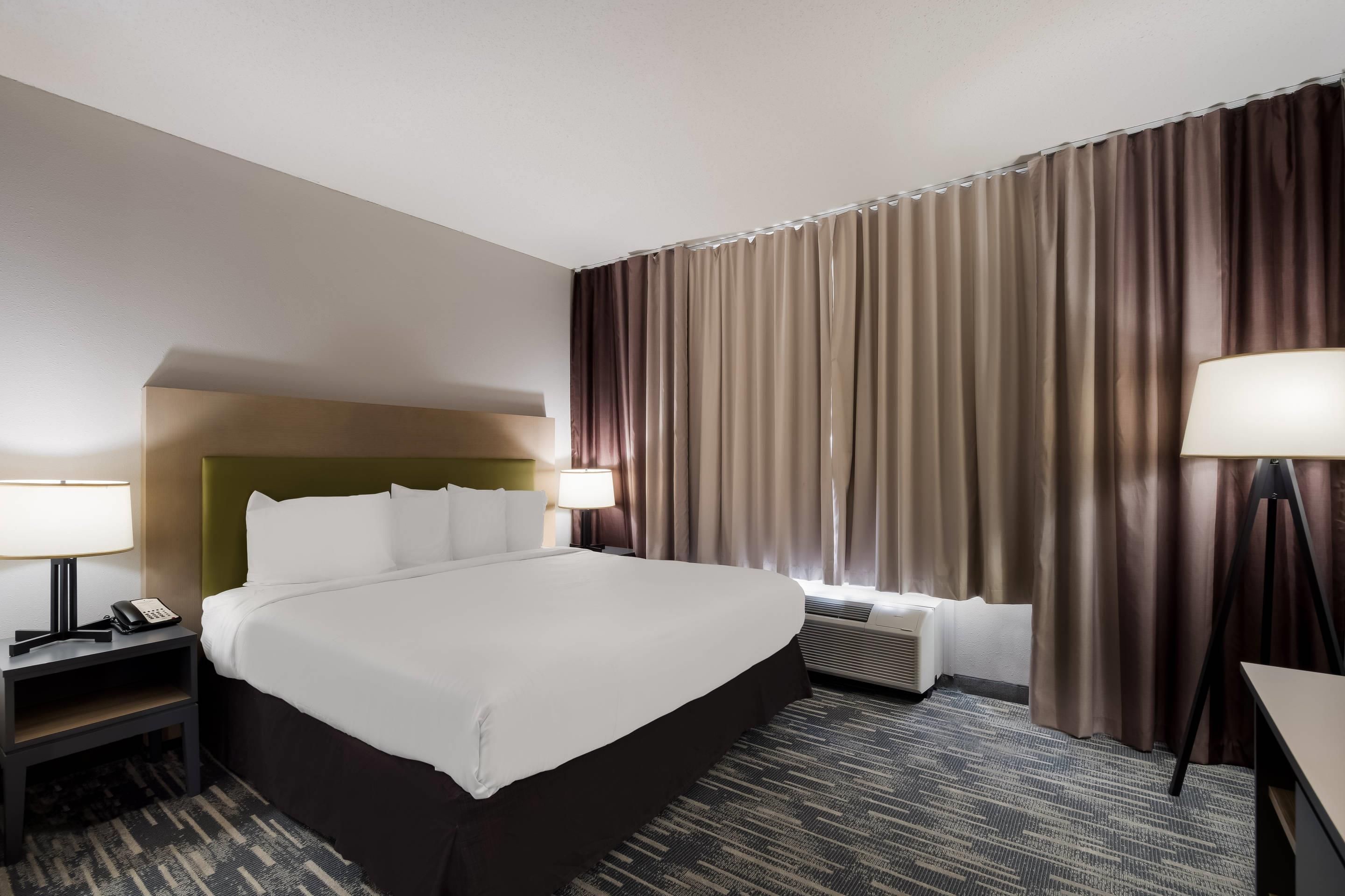 Een hotelkamer met een groot bed, twee nachtkastjes met lampen, een hoofdeinde, een raam met gordijnen en een vloerbedekking.