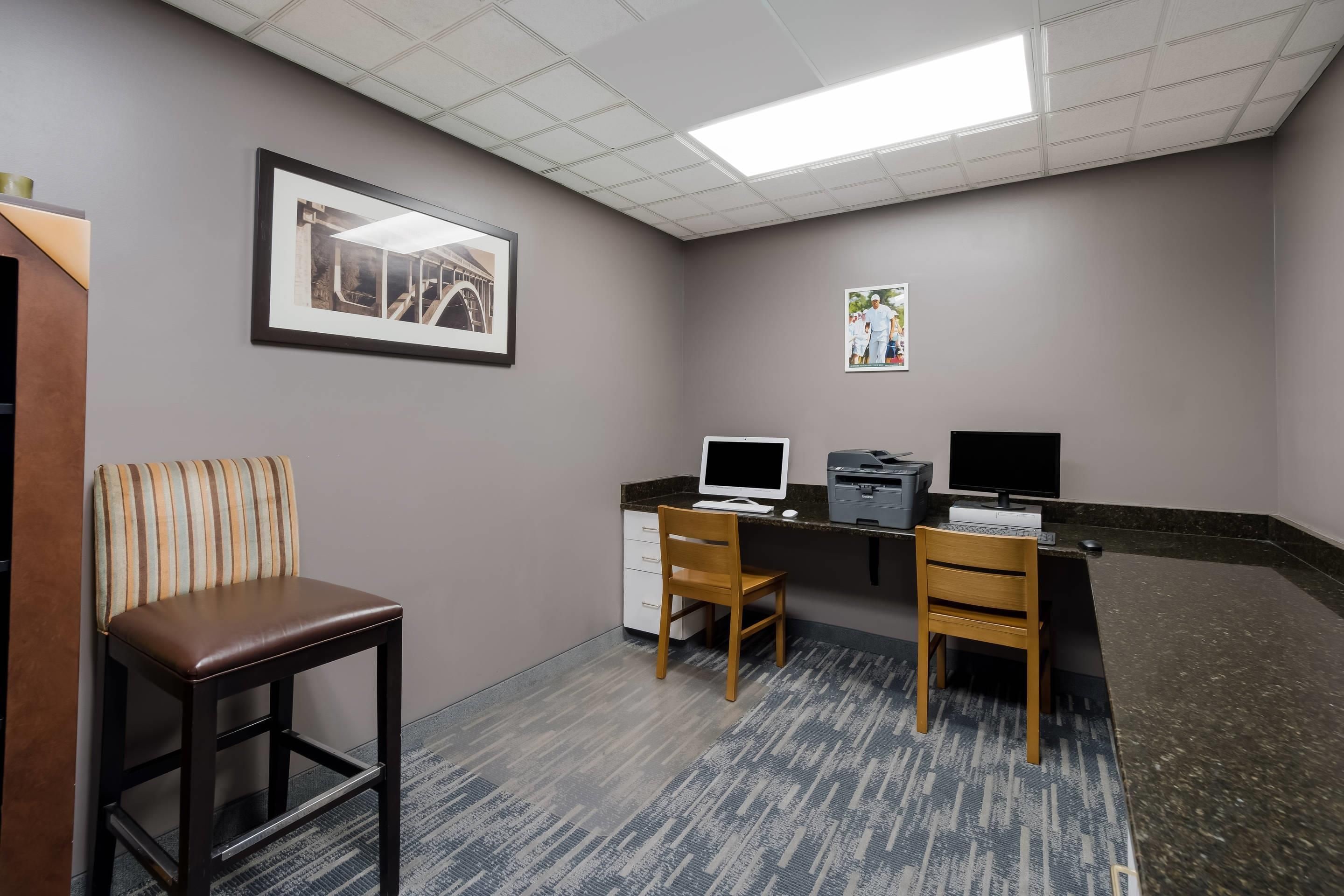 Ein Zimmer mit Schreibtisch, zwei Stühlen, einem Drucker, einem Computermonitor und gerahmten Bildern an der Wand.