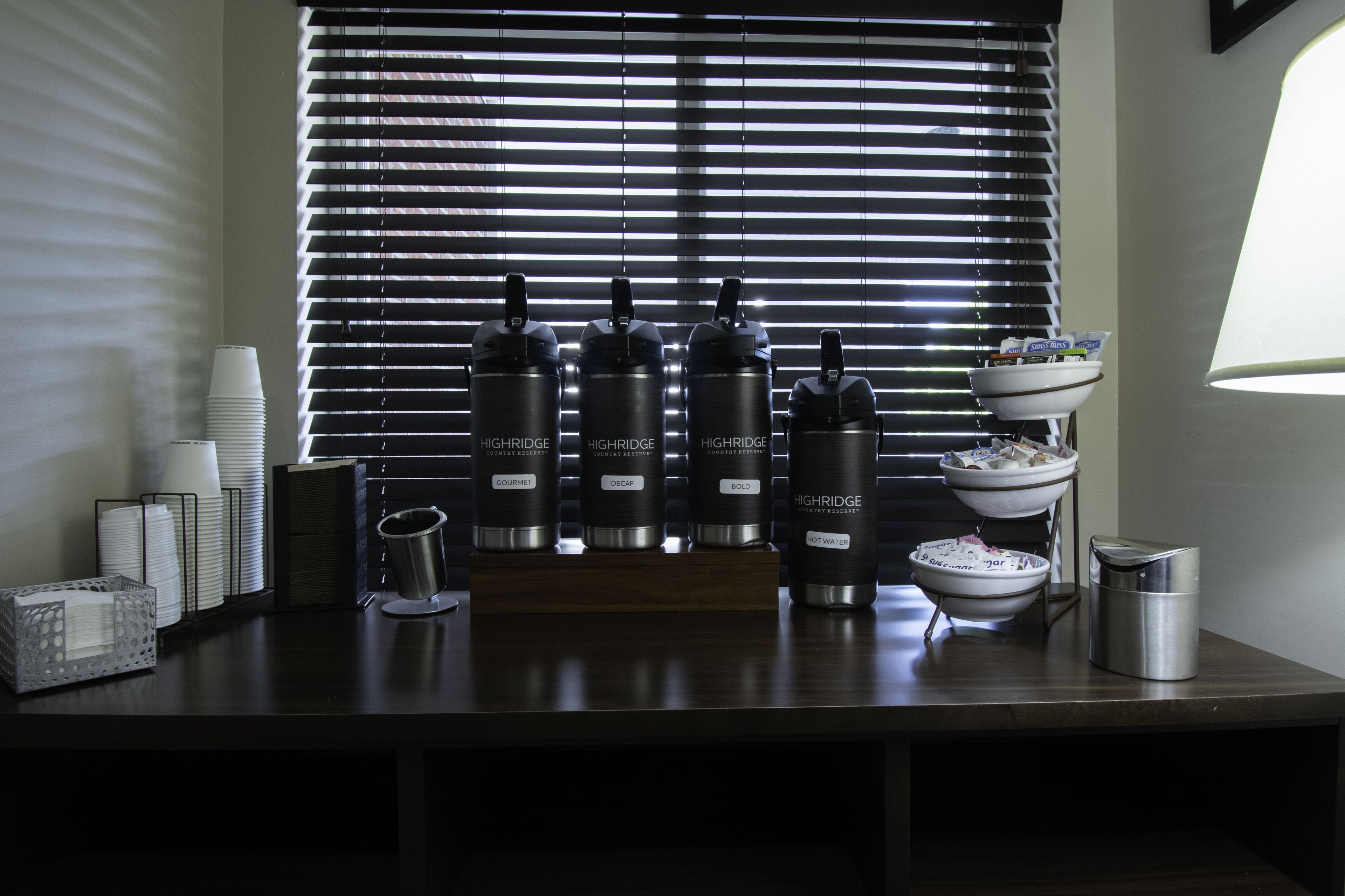 Una zona de bar con cuatro botellas de agua de la marca Highridge, etiquetadas como 'Gourmet', 'Descafeinado', 'Intenso' y 'Agua Caliente'. Hay vasos blancos apilados, una cesta metálica y un bol sobre un soporte.