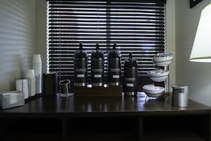 A bar area with four Highridge branded water bottles labeled 'Gourmet,' 'Decaf,' 'Bold,' and 'Hot Water.' There are stacked white cups, a metal basket, and a bowl on a stand. - Country Inn & Suites by Radisson, Augusta at I-20, GA (Augusta)