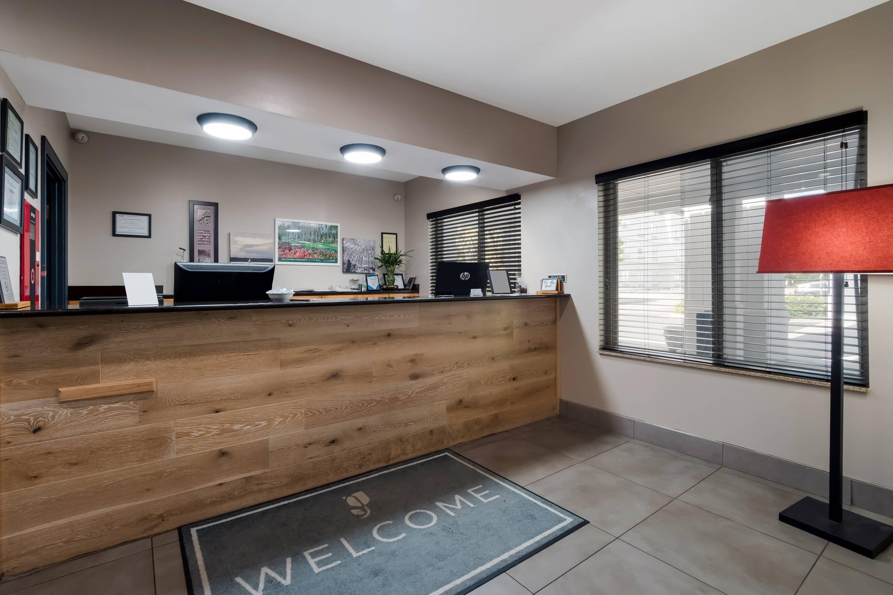 A reception area with a wooden reception desk, a 'WELCOME' mat, framed certificates, and a window with blinds.