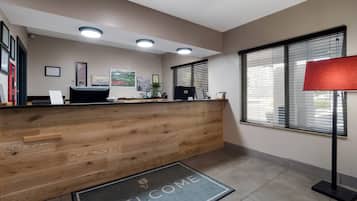 A reception area with a wooden reception desk, a 'WELCOME' mat, framed certificates, and a window with blinds.