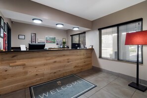 Et receptionsområde med en træreception, en velkomst måtte, indrammede certifikater og et vindue med persienner.