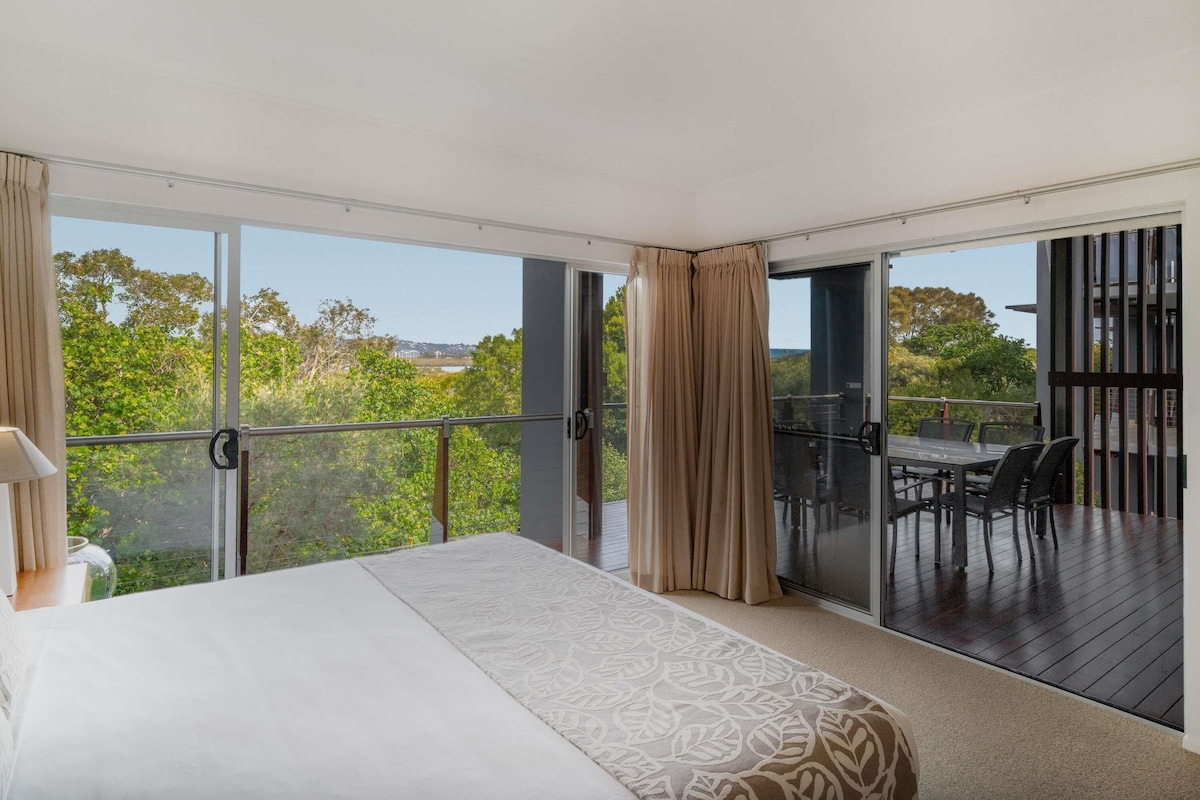 A bedroom with a large bed, a balcony with a table and chairs, and a view of greenery and buildings.