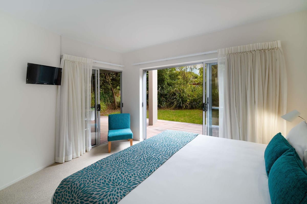 Een slaapkamer met een bed, een televisie, een stoel en uitzicht op een tuin via glazen deuren.