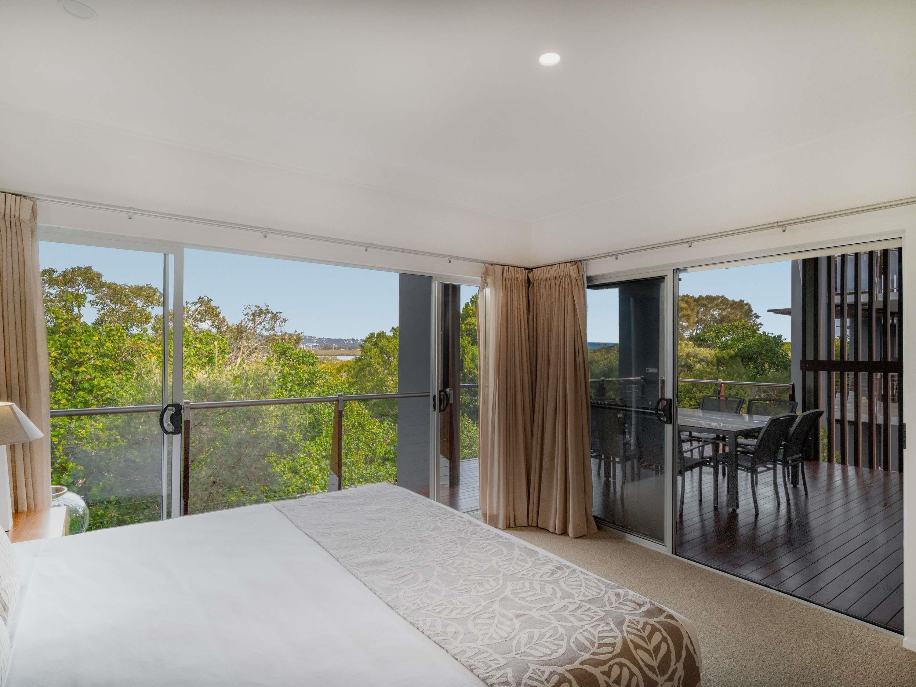 A bedroom with a large bed, a balcony with a table and chairs, and a view of greenery and buildings.