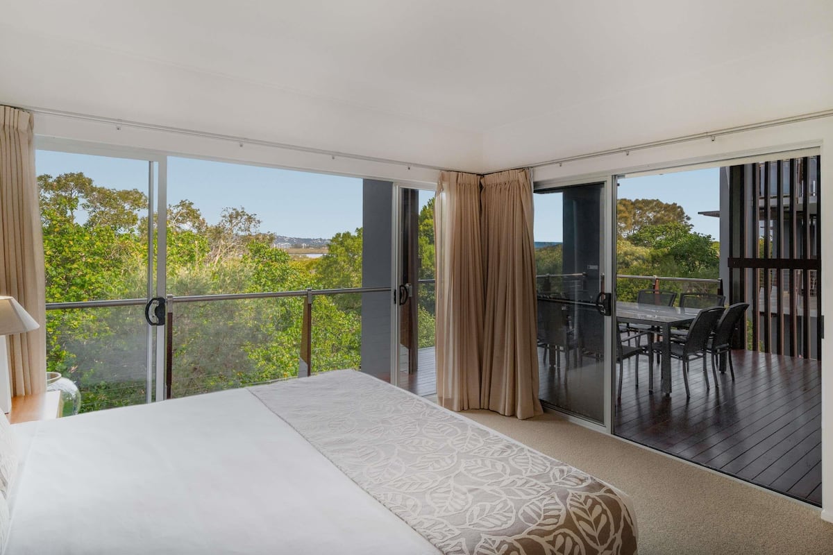 Ein Schlafzimmer mit einem großen Bett, einem Balkon mit Tisch und Stühlen sowie Blick auf Grünflächen und Gebäude.