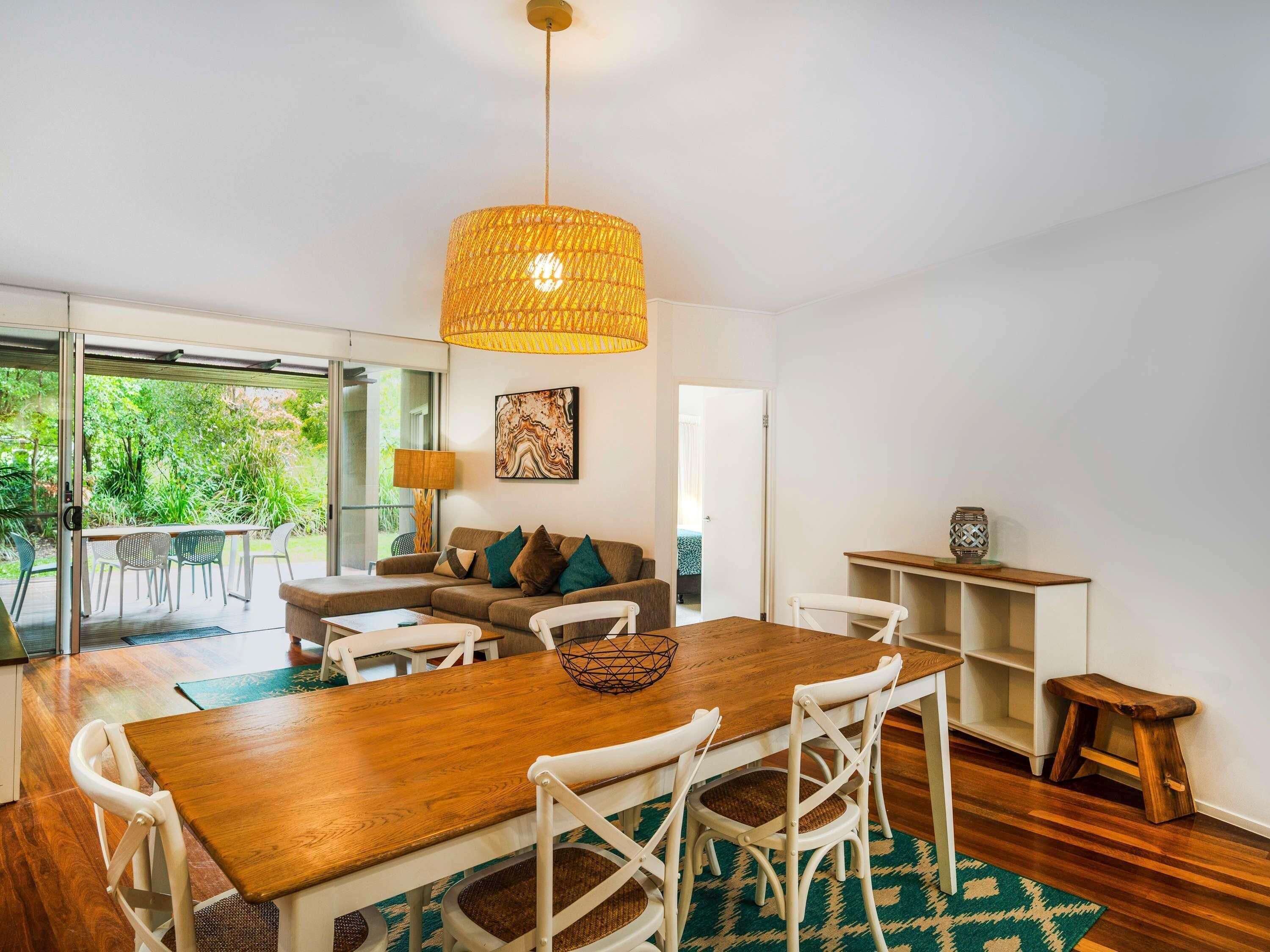 Una sala de estar moderna con una mesa de comedor de madera, sillas blancas, un sofá con cojines, una lámpara colgante y vistas a zonas verdes a través de puertas de vidrio.