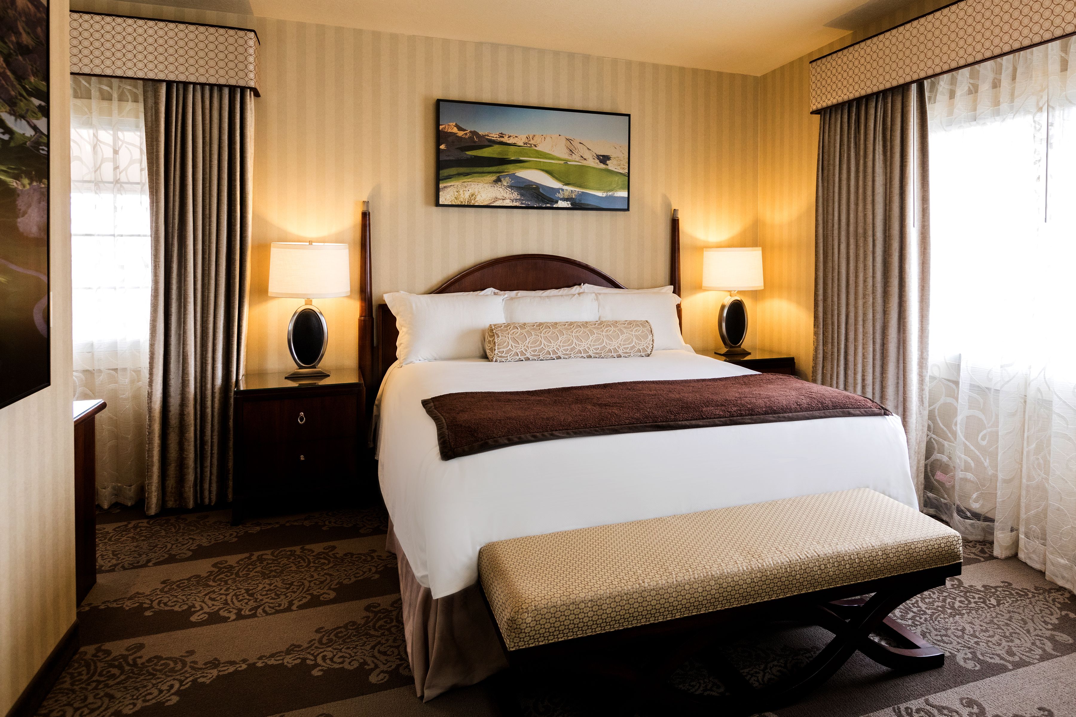 A hotel room with a large bed, two bedside lamps, a painting on the wall, and a patterned carpet.