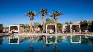 Una piscina con agua cristalina que refleja palmeras y edificios. Hay sillas de descanso y sombrillas alrededor de la piscina.