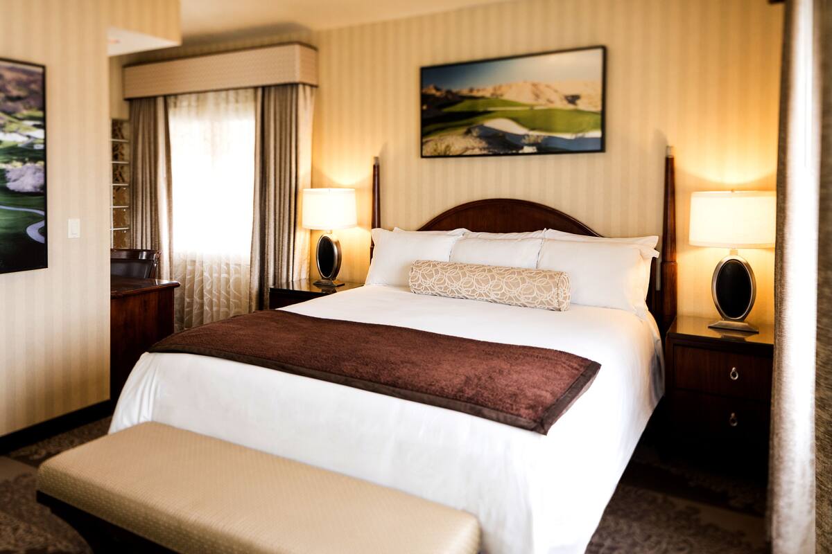 Une chambre d’hôtel avec un grand lit, deux tables de chevet avec des lampes, un banc au pied du lit et un tableau au mur.