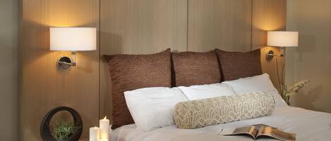 A neatly made bed with a book, two wall-mounted lamps, and a nightstand with a plant and candles.