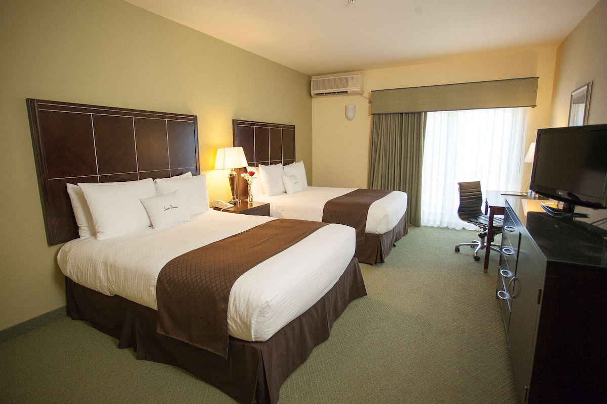 A hotel room with two beds, a desk with a chair, a television, and an air conditioning unit.