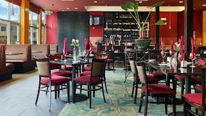 A restaurant with red upholstered booths, tables with red chairs, and a bar area with wine bottles. - DoubleTree by Hilton Napa Valley - American Canyon (American Canyon)