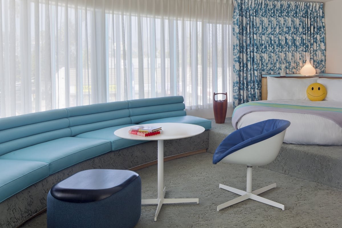 A modern hotel room with a blue sofa, a round white table, a blue chair, a bed with a smiley face pillow, and a patterned curtain.