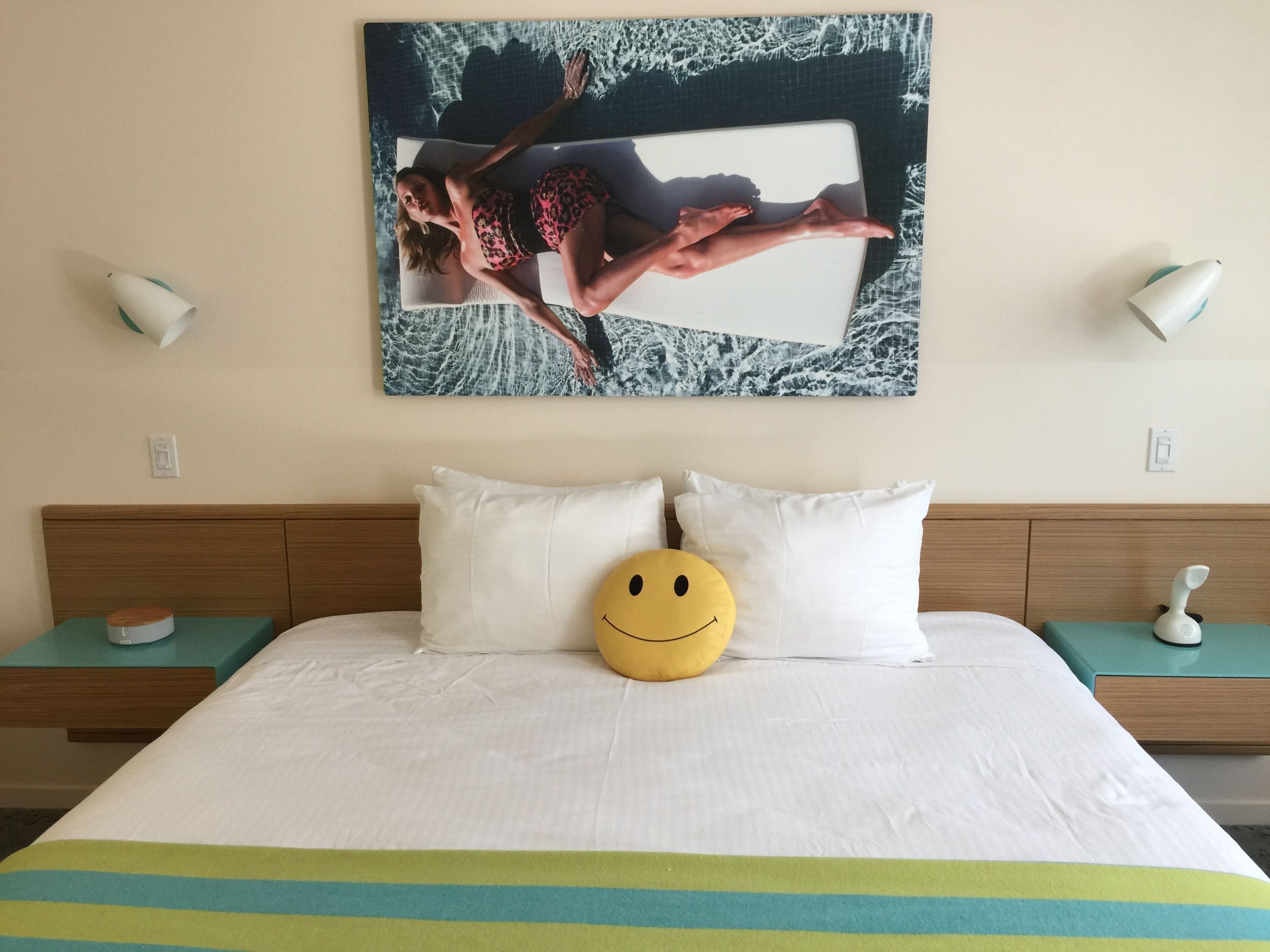 Quarto de hotel com uma cama, almofadas brancas, uma almofada em formato de rosto sorridente amarelo, um quadro de uma mulher de fato de banho e duas mesas de cabeceira com abajures.