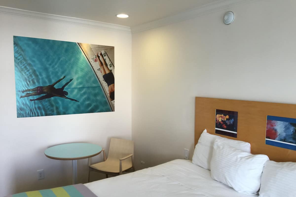 A hotel room with a bed, a small round table, a chair, and a painting of a swimmer.