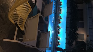 A pool area with chairs and a lounge chair, surrounded by a building with a blue-lit interior.