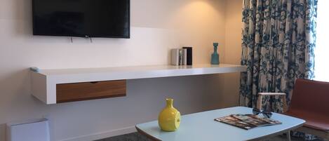 Un salon moderne avec un téléviseur à écran plat fixé au mur, une console blanche ornée d’un vase jaune, une petite table bleu clair et un fauteuil rouge.