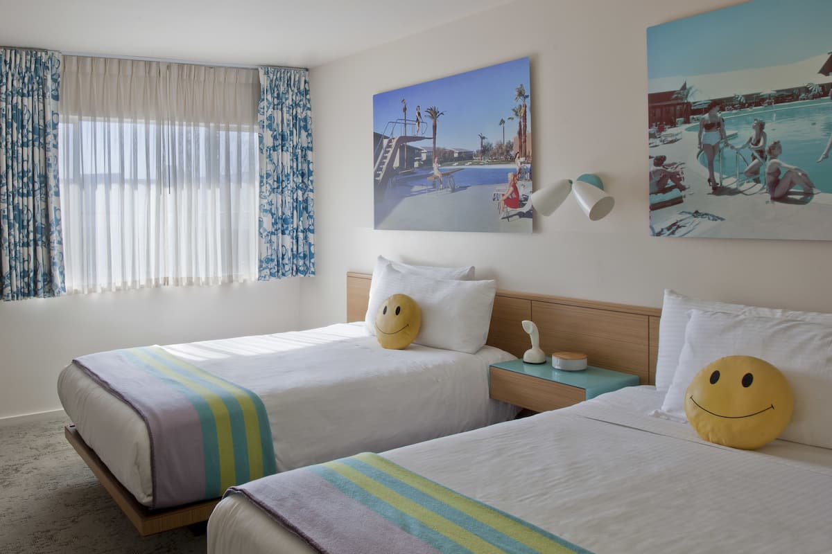 A hotel room with two beds, striped bedding, a smiley face pillow on each bed, a nightstand with a lamp, and a painting of a pool scene on the wall.