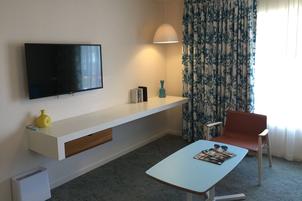 A modern hotel room with a flat-screen TV mounted on the wall, a white console table with a magazine, a small yellow vase, and a red chair.