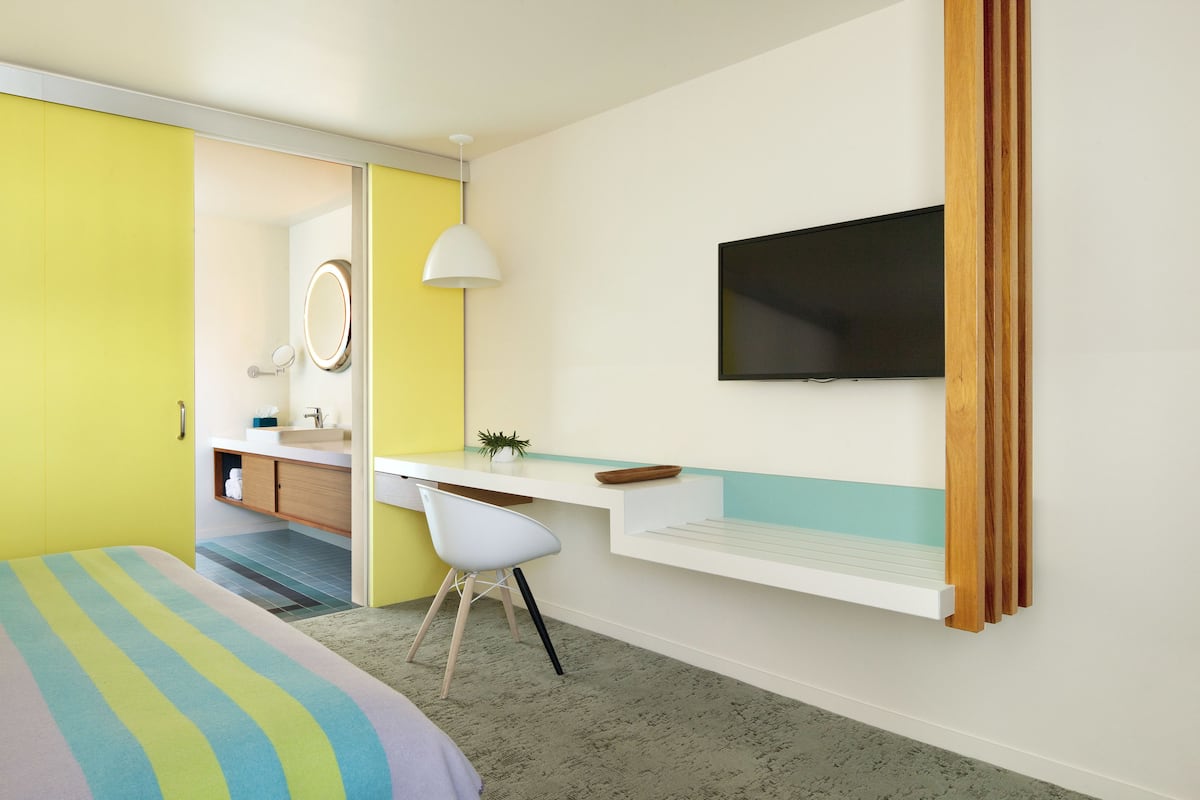 A modern hotel room with a bed, a desk, a chair, a TV, a mirror, and a bathroom visible through an open door.