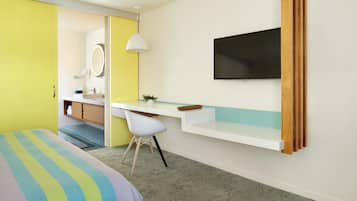 Quarto de hotel moderno com cama, escrivaninha, cadeira, televisão, espelho e banheiro visível através de uma porta aberta.