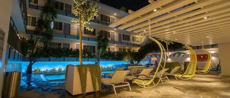 Un área junto a la piscina con sillones de descanso, una zona de estar cubierta y un edificio con balcones.