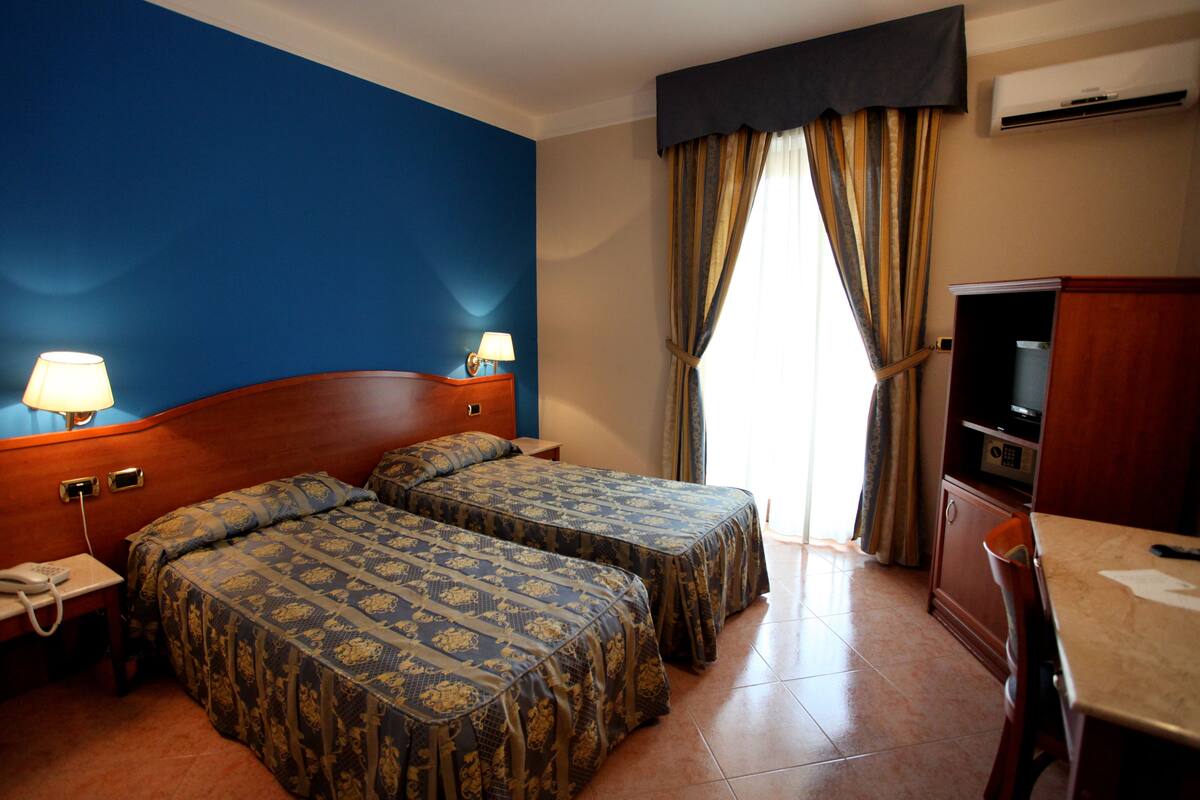 A hotel room with two beds, a desk with a lamp, a television, and an air conditioning unit.