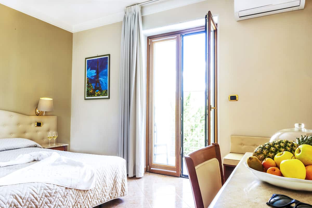 A hotel room with a bed, a bedside table with a lamp, a dining table with fruit, and a sliding glass door to the outside.