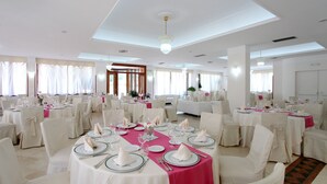 Salão de banquetes com mesas redondas preparadas para refeições, cadeiras brancas e um lustre central.