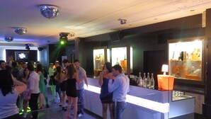 Uma área de bar com balcão, prateleiras com garrafas e luzes de discoteca no teto, e um grupo de pessoas em pé conversando.
