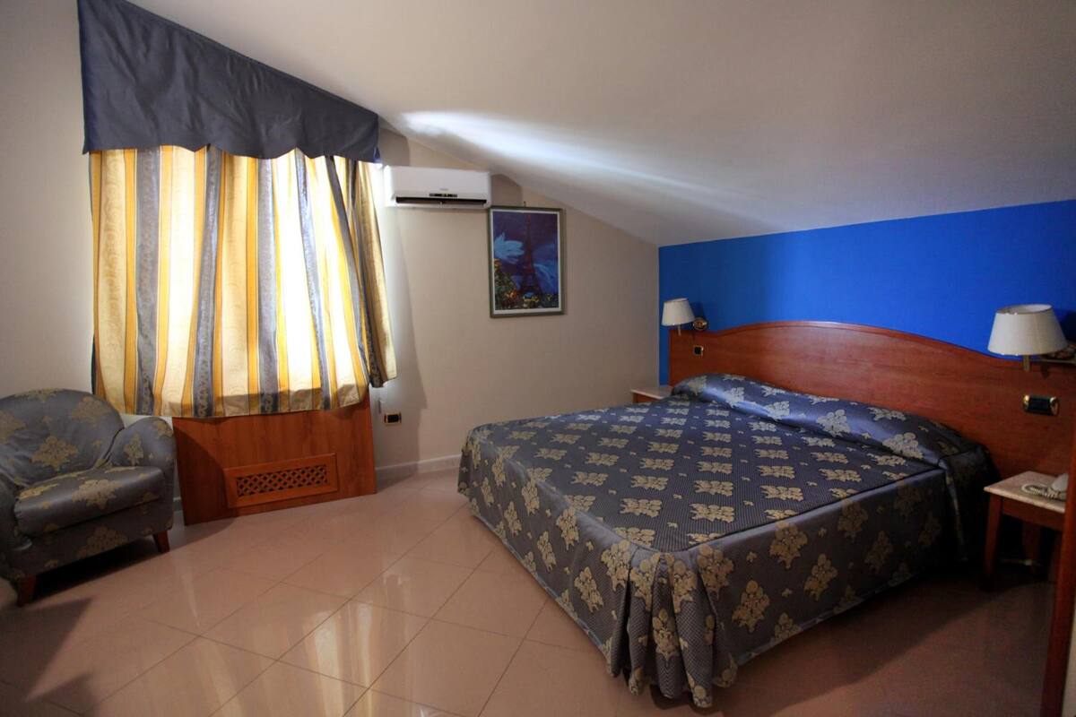 A hotel room with a bed, a chair, a nightstand, and a wall-mounted air conditioner.
