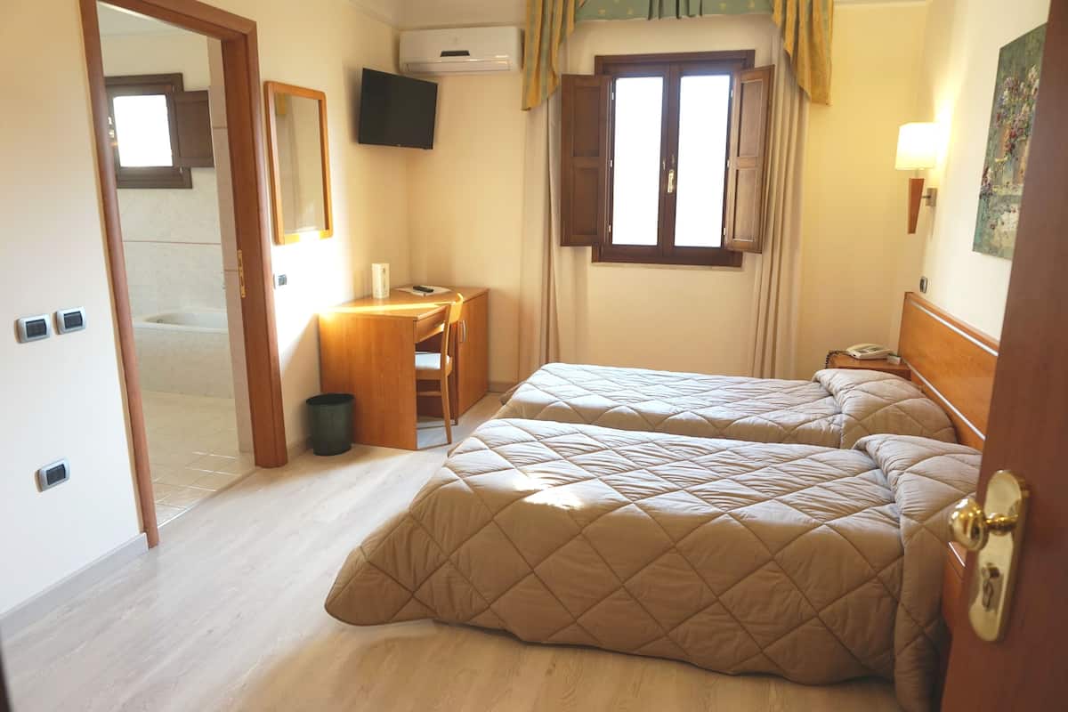 Een hotelkamer met twee bedden, een bureau, een stoel, een bijzettafeltje, een prullenbak, een raam met gordijnen, een tv aan de muur en een deur.