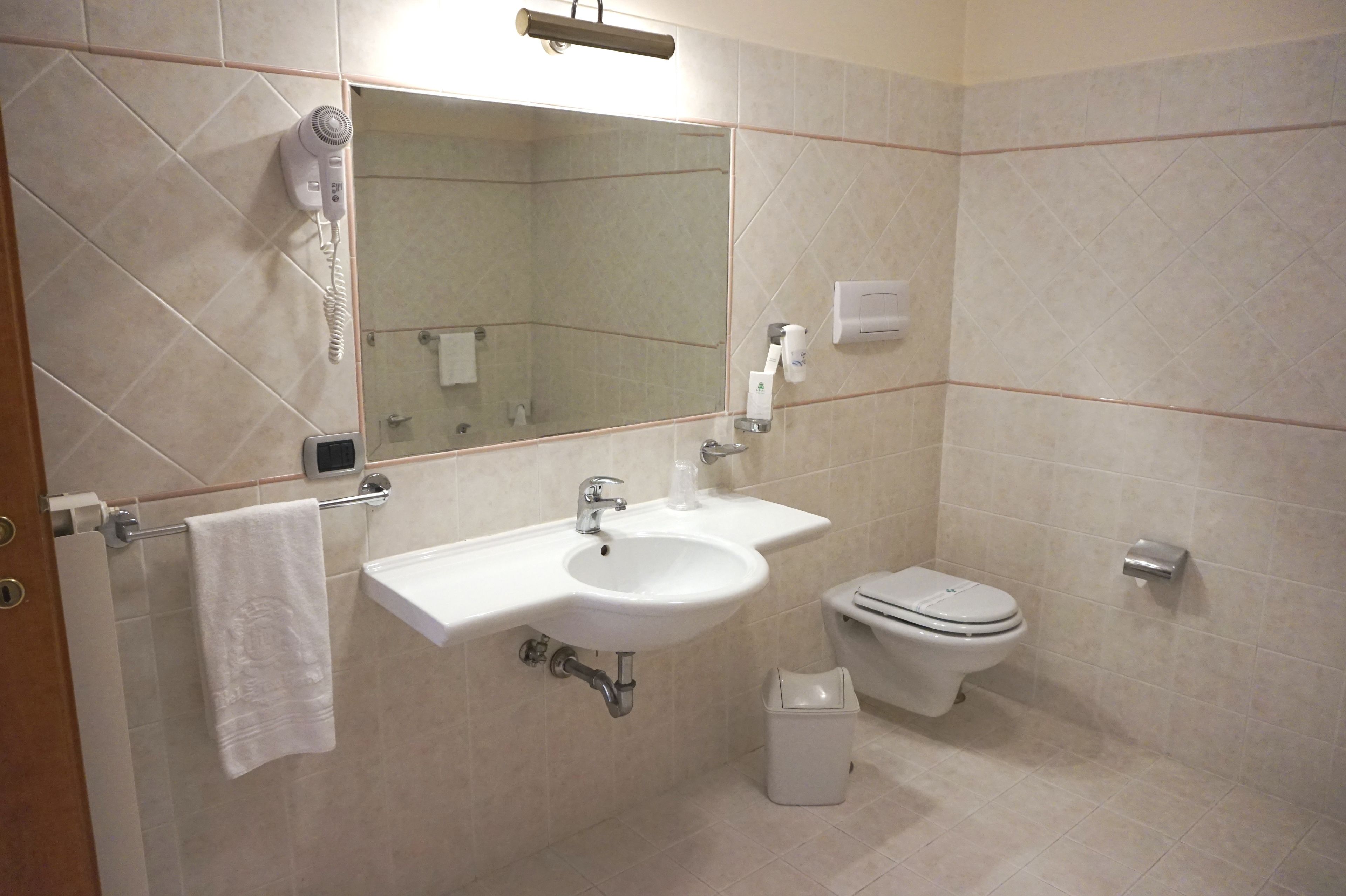 A bathroom with a sink, mirror, hairdryer, toilet, and trash bin.