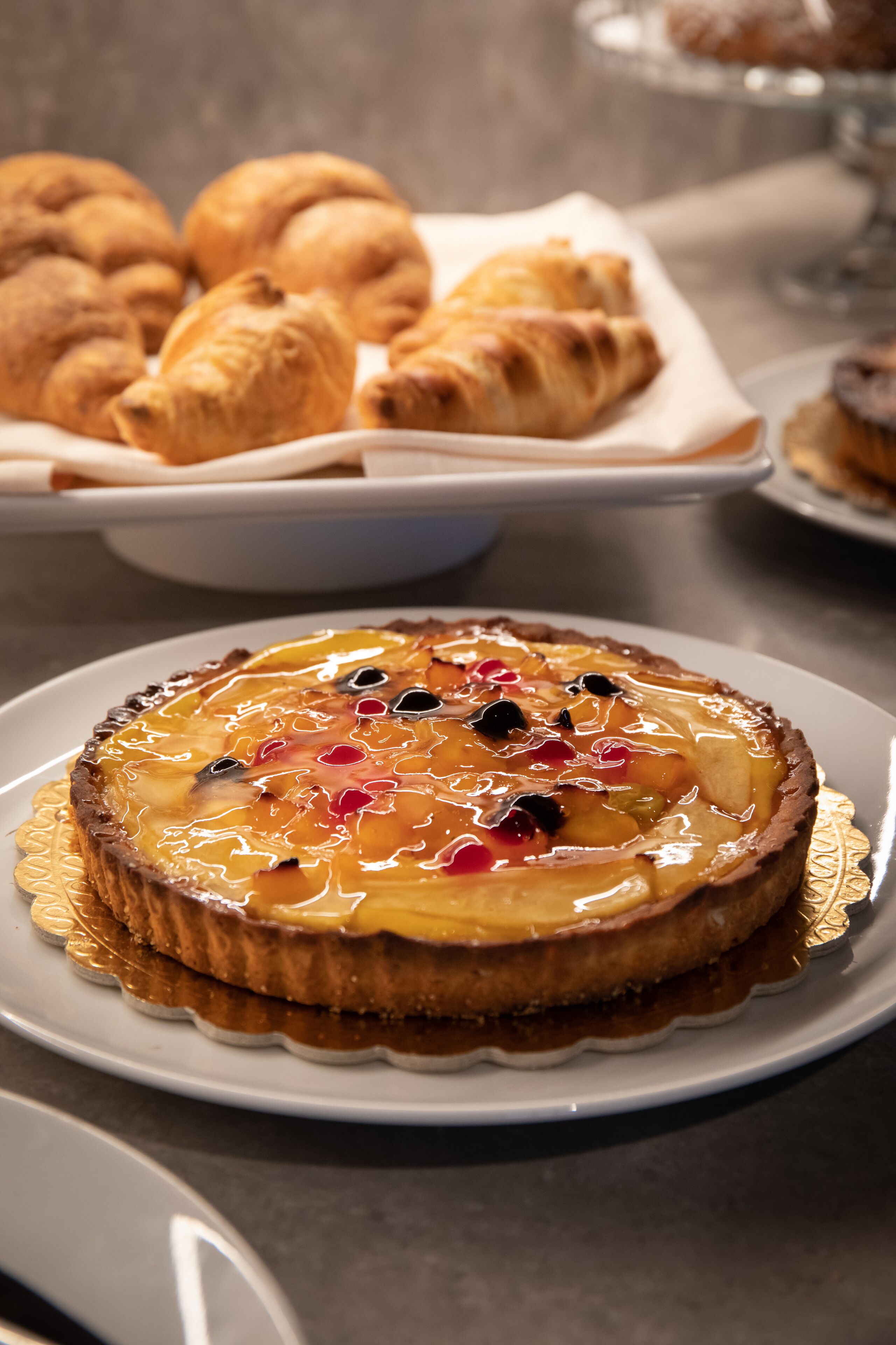 Un plato con una tarta cubierta de fruta y una bandeja con cruasanes al fondo.