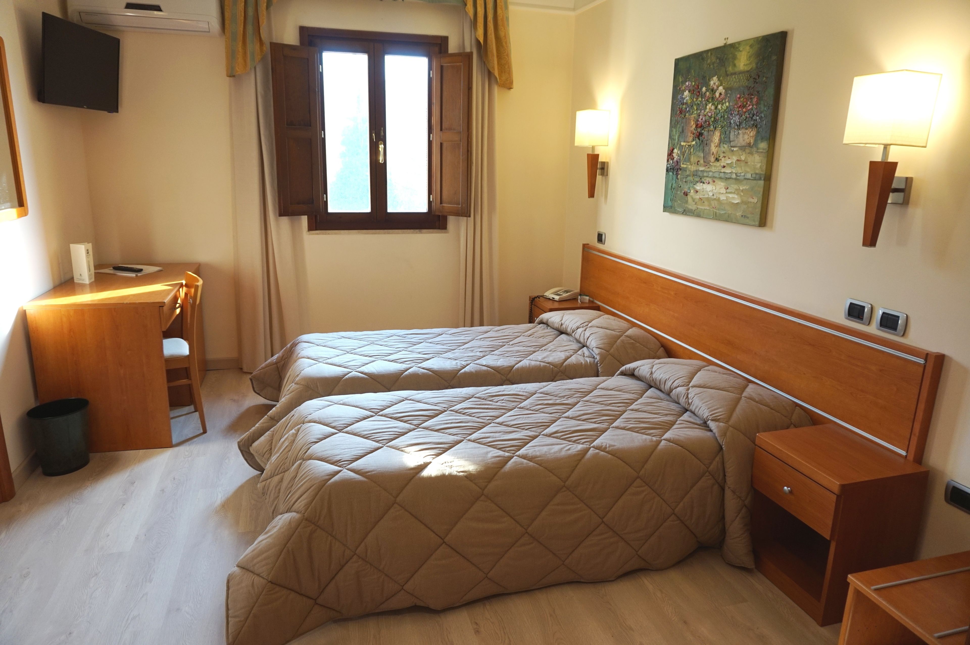 Habitación de hotel con dos camas, un escritorio, una silla, un cubo de basura, una mesita de noche, una lámpara y un cuadro en la pared.