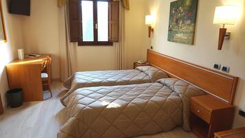Habitación de hotel con dos camas, un escritorio, una silla, un cubo de basura, una mesita de noche, una lámpara y un cuadro en la pared.
