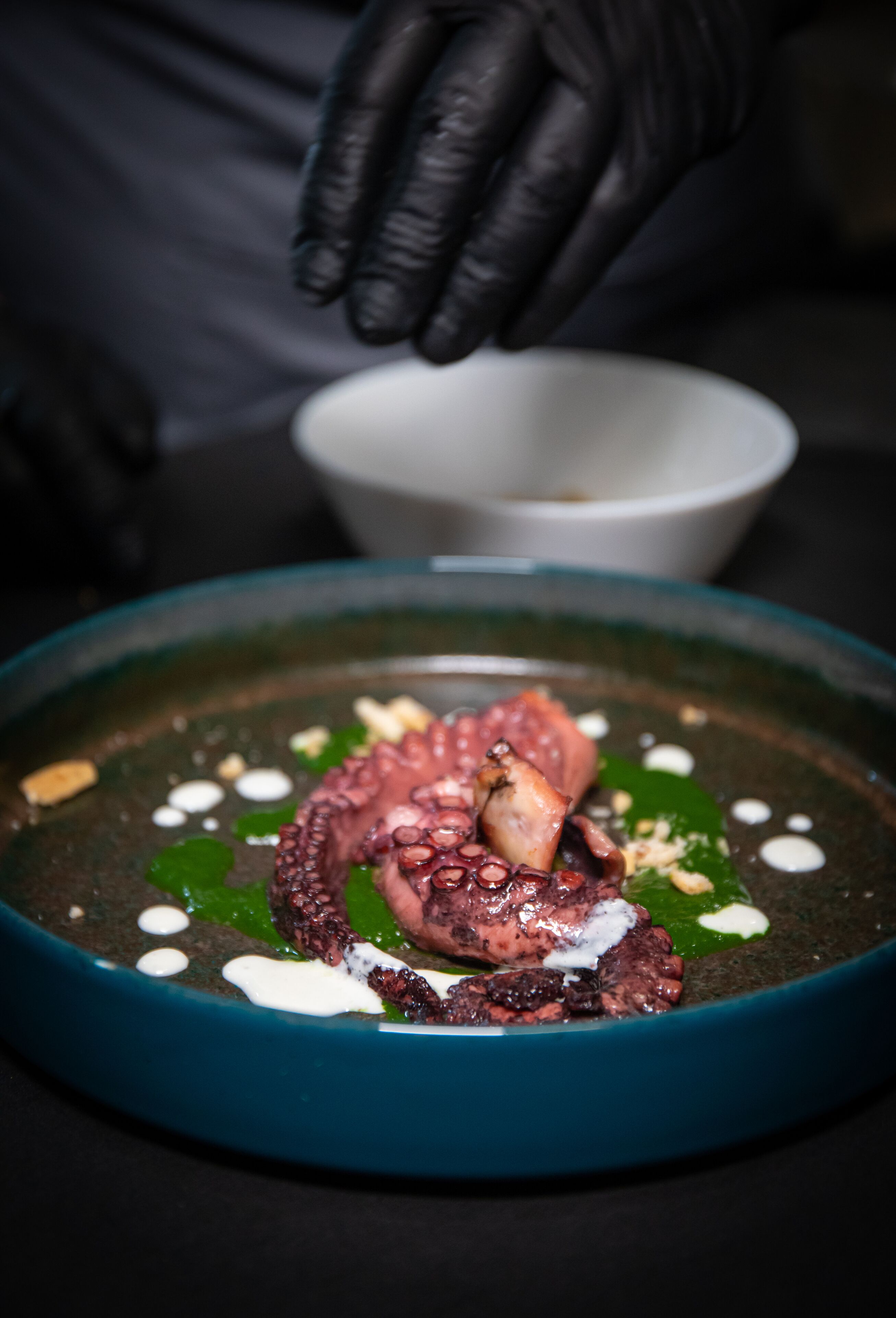 A person wearing black gloves preparing a dish with octopus and garnishes in a teal bowl.