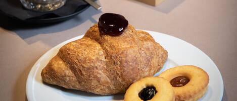 Desayuno con un croissant, dos galletas pequeñas, una taza de café y una caja de crema de chocolate sobre una mesa.