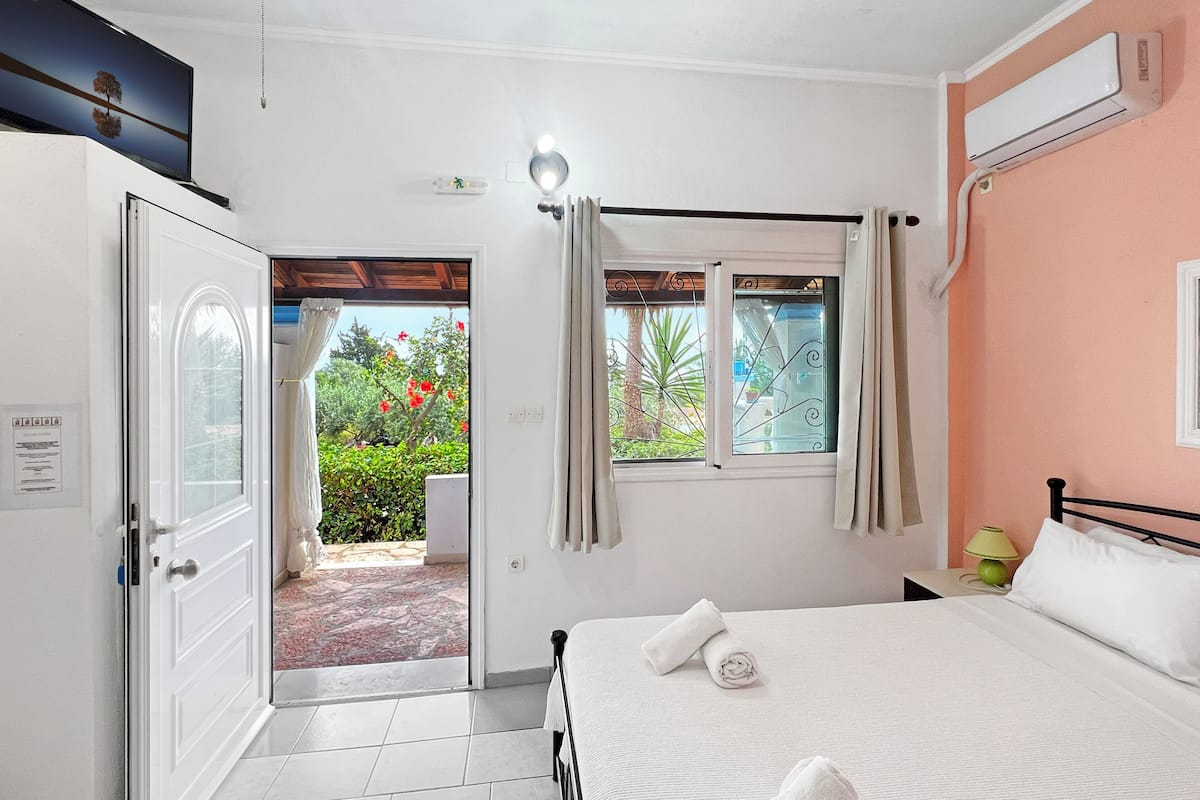 A bedroom with a bed, a window with curtains, a door, and a view of a garden.