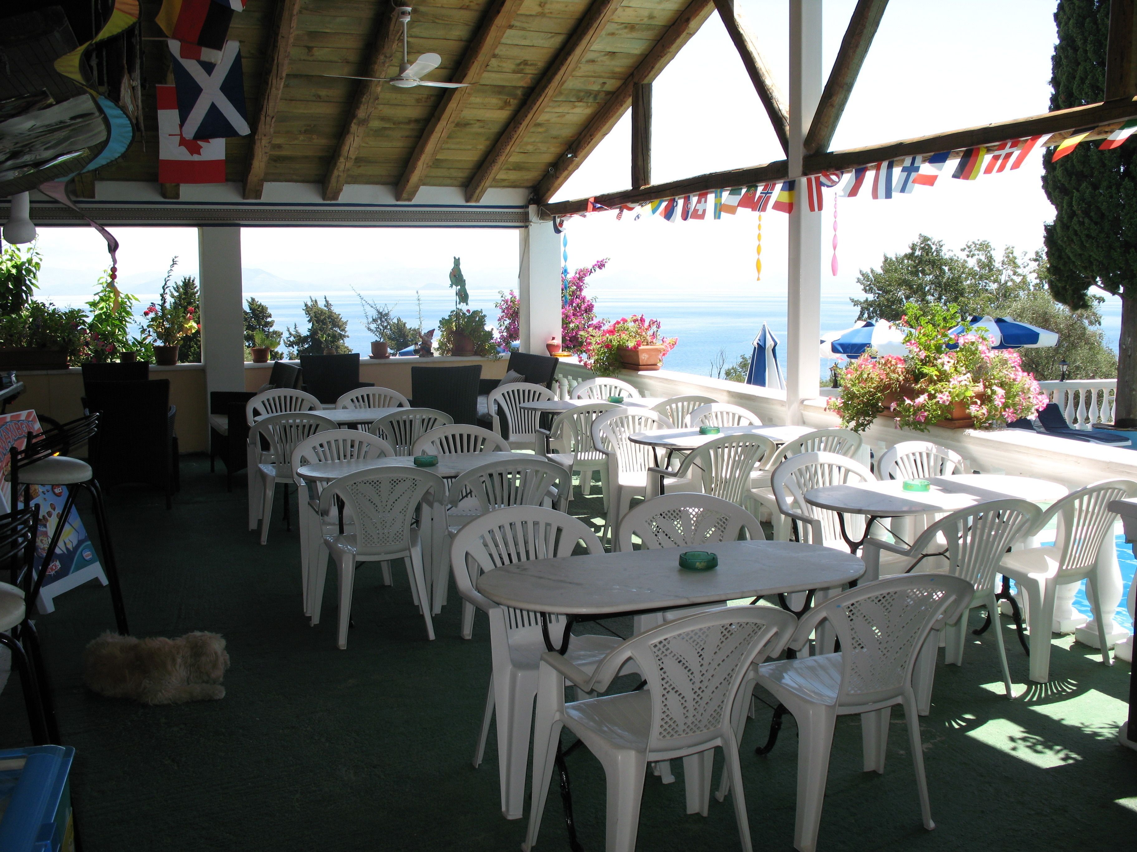 Une terrasse extérieure avec des chaises et des tables en plastique blanc, des plantes en pot et une vue sur la mer.