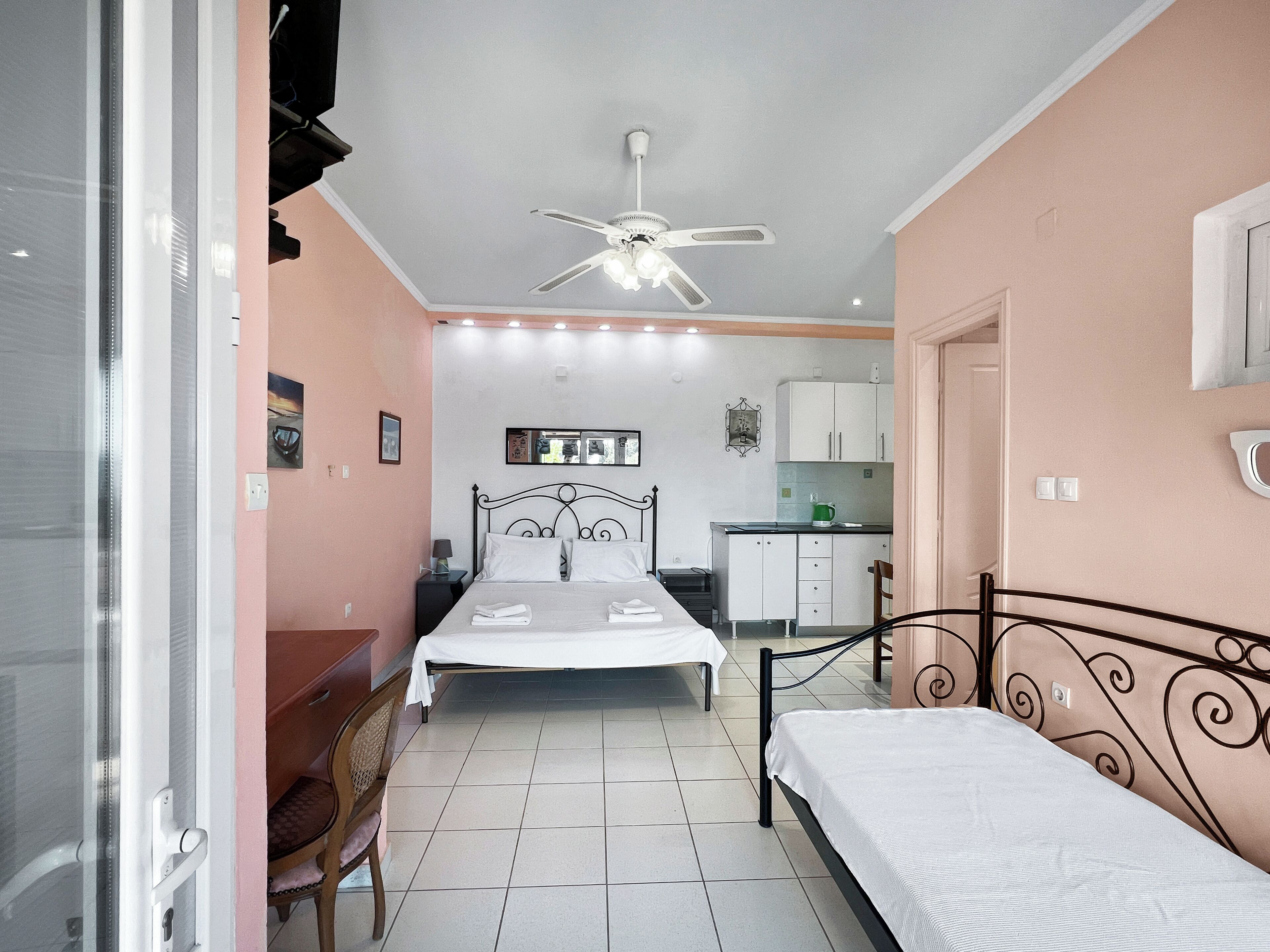 A bedroom with a bed, a desk, a chair, a kitchenette, and a ceiling fan.